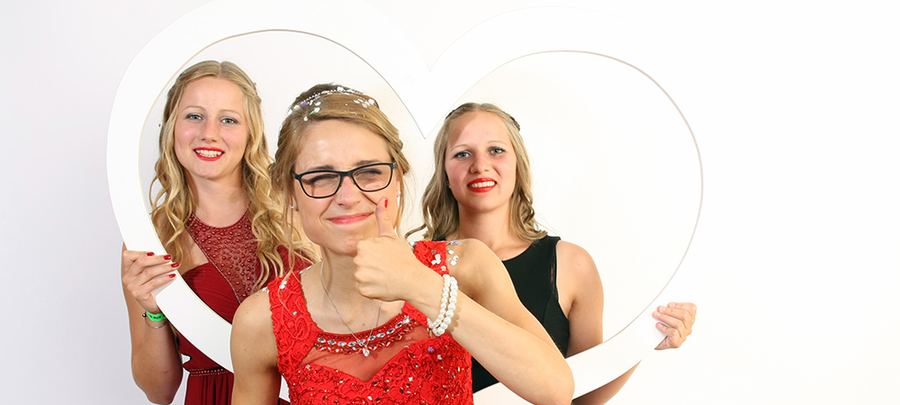 Drei junge Frauen in Abendkleidern posieren mit einem herzförmigen Fotorahmen beim Abiball